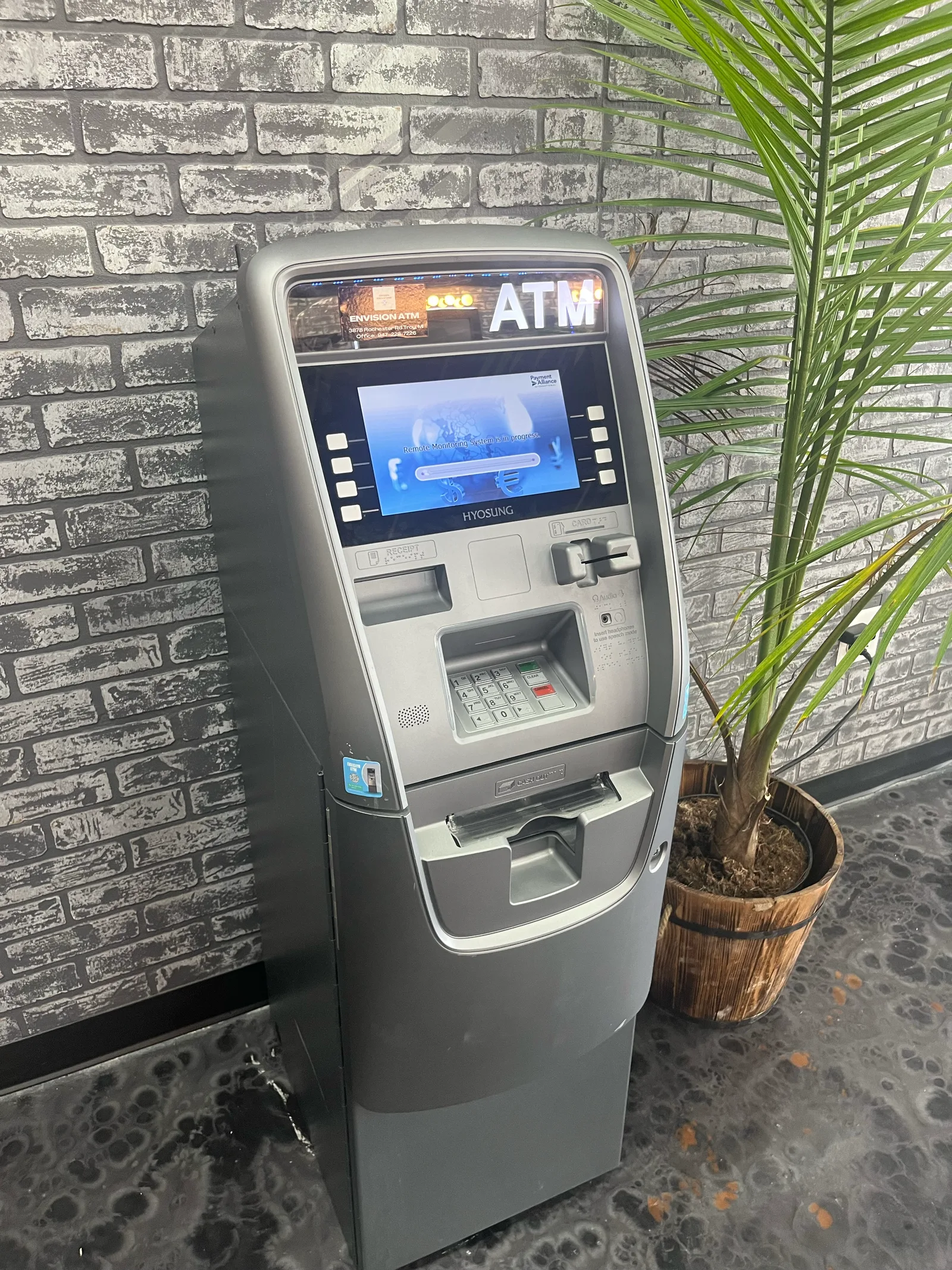 An Envision ATM machine installed against a grey brick wall beside a potted palm